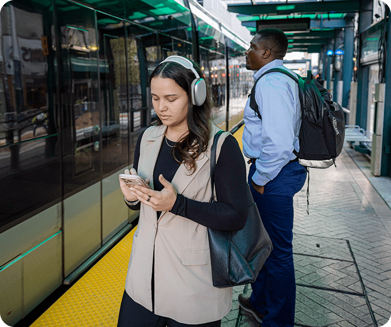 Two people wait for a light rail train. One person looks at their phone while another one looks at the train doors.